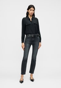 Femeie purtând un tricou negru cu mâneci lungi, curea neagră cu ținte, blugi gri închis și balerini negri cu vârf ascuțit, stând în fața unui fundal simplu.