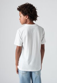 Bílý bavlněný tričko s krátkým rukávem, kulatým výstřihem a malým logem na zadní straně. Nosené s světle modrými džíny. Jasná textura.