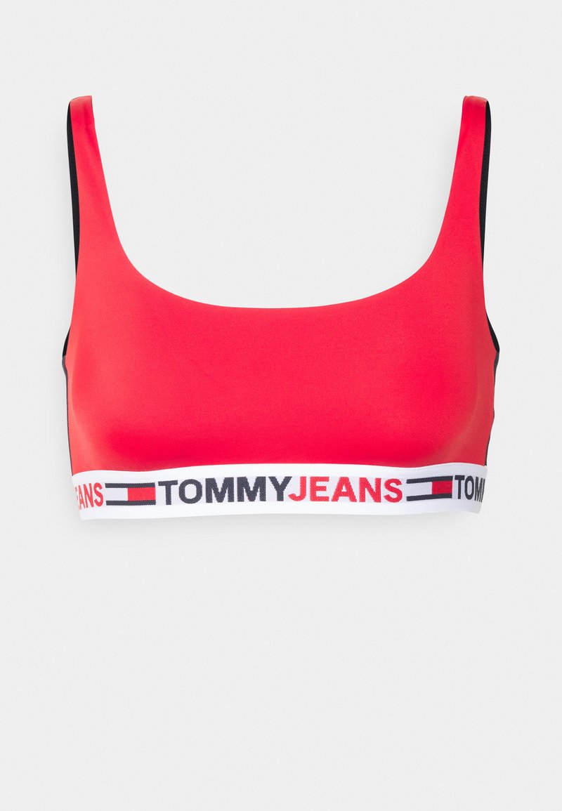 Sutiã desportivo vermelho com alças largas, borda preta e uma faixa elástica branca com a marca "TOMMY JEANS" em letras garrafais.
