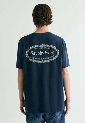 Navyblauw katoenen T-shirt met een grote witte ovale afbeelding op de achterkant, voorzien van tekst en decoratieve touwaccenten. Korte mouwen, ontspannen pasvorm.