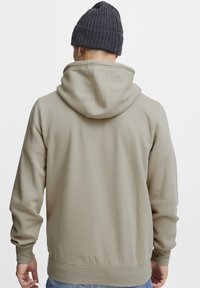 Sudadera con capucha color caqui de textura suave, con puños y dobladillo acanalados. Combina con un gorro de punto oscuro, mostrando un diseño casual.