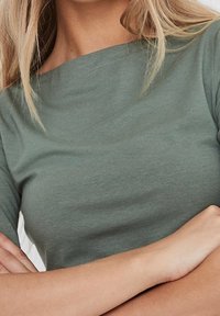 Svetle olivově zelený top s krátkým rukávem, širokým výstřihem a uvolněným střihem. Měkká, hladká bavlněná tkanina s mírným elastickým efektem. Jednoduchý design, bez viditelných vzorů.