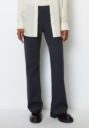 Person iført sorte bukser med vide ben, sorte sko med chunky såler og en off-white skjorte med knapper og synlige manchetter.