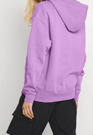 Person trägt einen lavendelfarbenen Hoodie mit Kapuze und schwarze Cargohosen, von hinten vor einem einfarbigen hellen Hintergrund gezeigt.