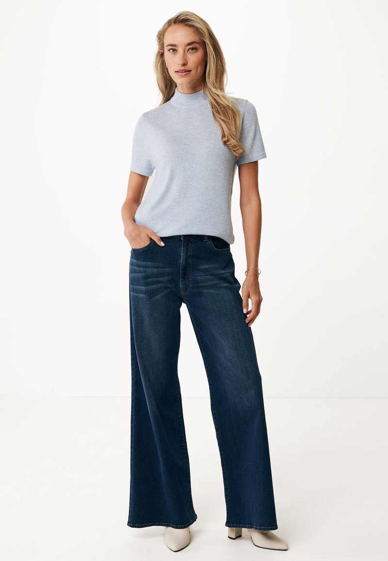 Kobieta z długimi blond włosami, ubrana w jasnoniebieską krótką bluzkę z dzianiny, ciemne szerokie dżinsy, białe czółenka, stojąca z jedną ręką w kieszeni.