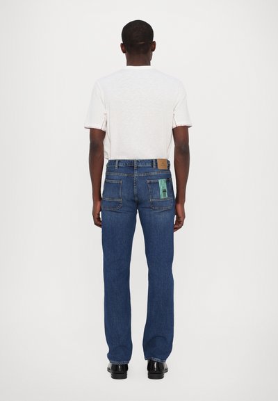 Jeans en denim bleu avec jambes droites, cinq poches et un patch sur la ceinture ; présente un design distinctif à rayures vertes sur la poche arrière.