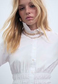 Donna bionda con capelli sciolti indossa una camicia bianca con bottoni e vita elastica, accompagnata da collane di catene d'oro sovrapposte, su uno sfondo chiaro e uniforme.