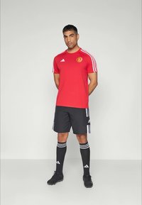 Piros adidas sportpóló fehér csíkokkal, Manchester United logóval, fekete rövidnadrággal, fekete zoknival és focicipővel párosítva.