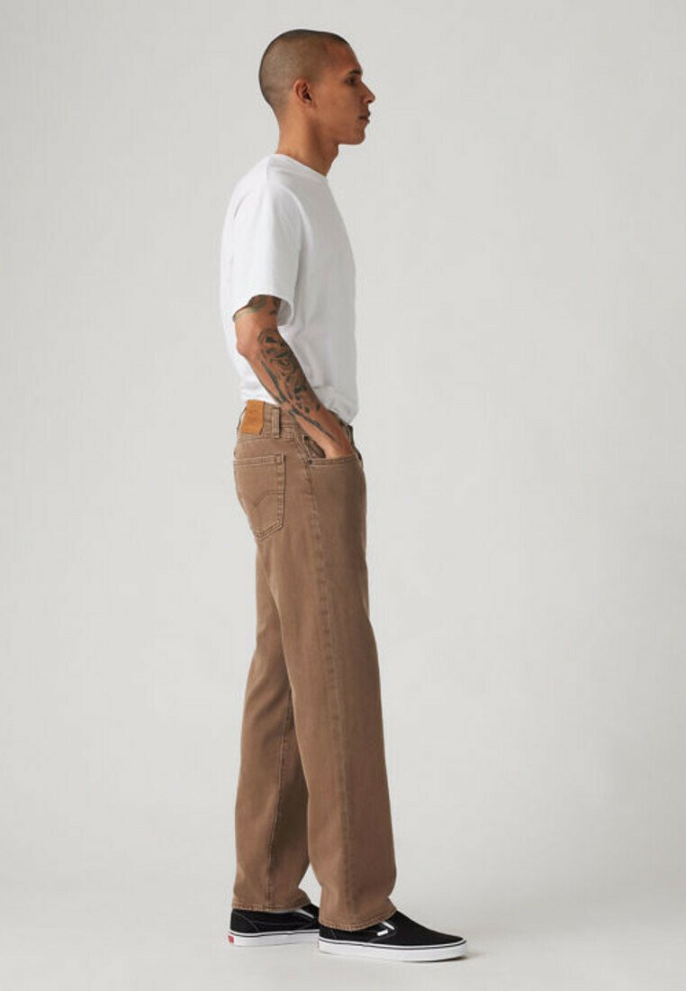 Man with tattooed arm stands sideways wearing white t-shirt, brown jeans, and black slip-on shoes against plain light background.