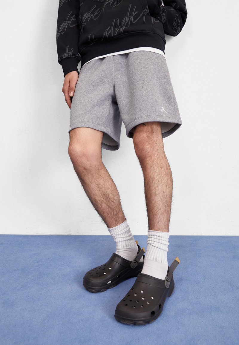 Grijze sportshorts met een subtiele logo, gecombineerd met zwarte instapklompen en witte geribde crew-sokken, tegen een blauwe ondergrond.