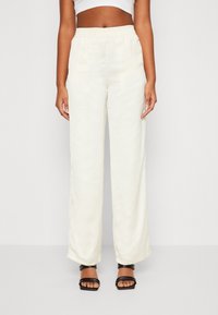 Vrouw in beige hoge taille wijde broek met elastische tailleband, gecombineerd met zwarte hoge hakken sandaaltjes en een witte crop top.