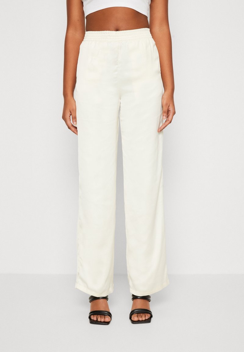 Vrouw in beige hoge taille wijde broek met elastische tailleband, gecombineerd met zwarte hoge hakken sandaaltjes en een witte crop top.