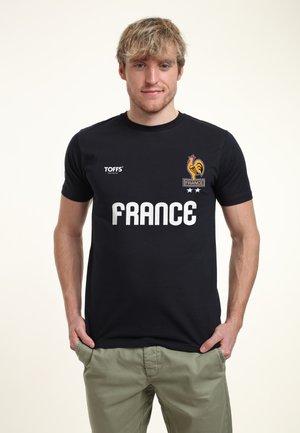 Junger Mann mit blondem Haar, der ein schwarzes Frankreich-Shirt mit einem Hahn-Logo und weißem Text trägt, steht mit Händen in den Taschen vor einem schlichten Hintergrund.