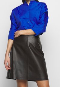 Chemise boutonnée en satin bleu avec un col haut et des manches trois-quarts, associée à une jupe trapèze en cuir noir.