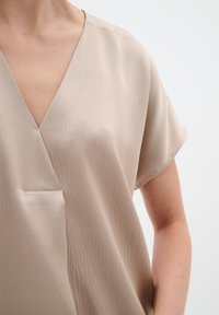 Beige lyhythihainen paita V-pääntiellä, jossa on teksturoitu kangas ja etutasku. Suunnittelu on väljä ja minimalistinen.