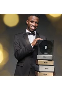 Zwarte tuxedo met een vlinderdasje toont een zwarte doos met het label "BOSS," gestapeld op gouden en zilveren dozen, met scherpe logo's. Zachte achtergrond in onscherpe focus.