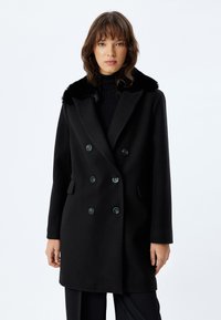 Cappotto di lana nero con design doppiopetto, colletto in pelliccia sintetica e due tasche frontali. Dotato di grandi bottoni neri e spalle strutturate.