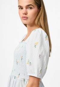 Gaiši zils izšūts kleitas modelis ar smocked bodi un pūkainām piedurknēm, ar dzeltenu un purpura ziedu rakstiem uz teksturētas auduma.