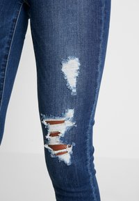 Närbild av mörkblå denimjeans med slitna, fransiga hål som avslöjar huden på låret mot en neutral bakgrund.