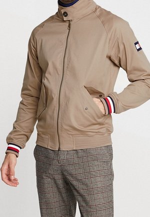 Mann trägt eine beige Zip-up-Jacke mit gestreiften Bündchen und karierte graue Hose, eine Hand in der Tasche, steht vor einem schlichten Hintergrund.