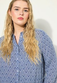 Camicia blu a stampa con disegni floreali bianchi, maniche lunghe, chiusura frontale con bottoni e colletto. Indossata con una vestibilità ampia e una texture di tessuto morbida.