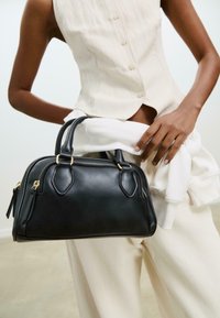 Sac à main en cuir noir de forme arrondie, avec deux poignées supérieures et des finitions en or. Il présente des coutures soignées et deux compartiments zippés.