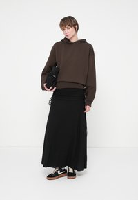 Hnedá mikina s kapucňou, čierna maxi sukňa a čierne tenisky s bielymi akcentmi. Model drží malú čiernu tašku. Minimalistický dizajn.