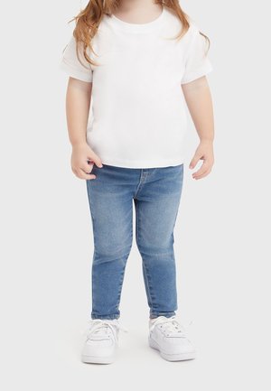 Niño pequeño con camiseta blanca de manga corta, vaqueros ajustados azules y zapatillas blancas, de pie contra un fondo claro y sencillo.