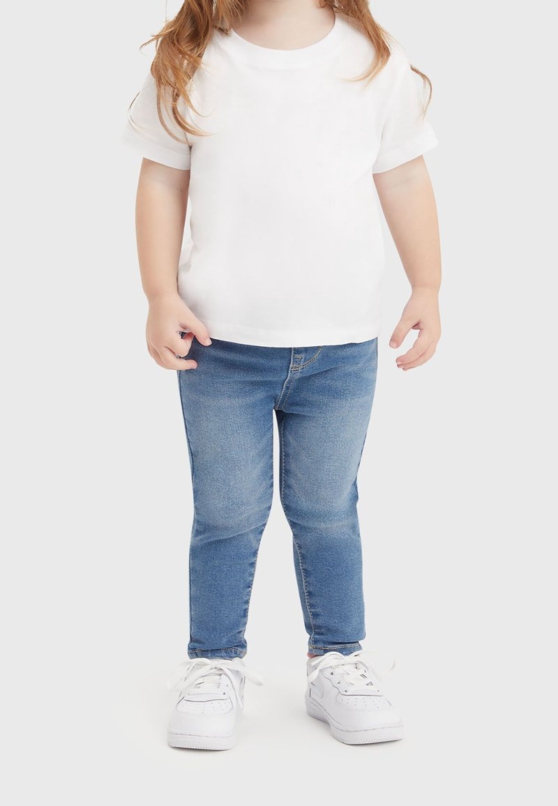 Tout-petit portant un t-shirt blanc à manches courtes, un jean skinny bleu et des baskets blanches, debout devant un fond clair uni.