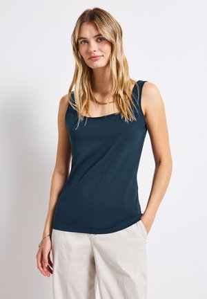 Top senza maniche di color teal scuro con scollatura quadrata e spalline spesse, realizzato in tessuto morbido e liscio. Abbinato a pantaloni di colore chiaro.