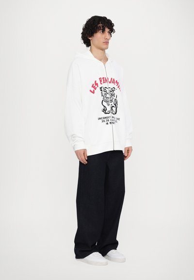 Λευκό φούτερ με φερμούρ σε στυλ ζακέτας με γραφικό σχέδιο σε κόκκινο και μαύρο, συνδυασμένο με σκούρα μπλε χαλαρά παντελόνια και λευκά αθλητικά παπούτσια.