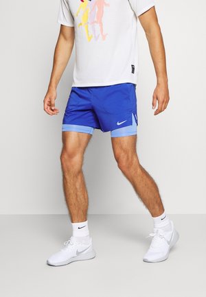 Homme portant un short de course Nike bleu superposé, un t-shirt blanc à motif, des chaussettes blanches Nike et des chaussures de course blanches Nike.