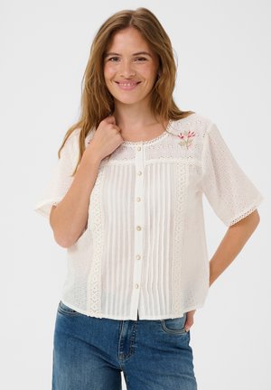 Donna sorridente, indossa una camicetta bianca a maniche corte con pizzo, bottoni e ricamo floreale, abbinata a jeans di denim blu, sfondo chiaro.
