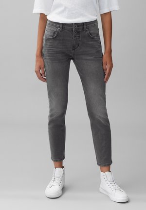 Personne portant un jean slim noir délavé et des baskets blanches montantes debout devant un fond gris.