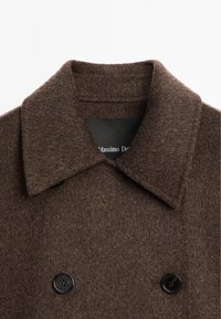 Brun uldfrakke med en bred krave, dobbeltradet foran og sorte knapper. Blød tekstur med et klassisk design.