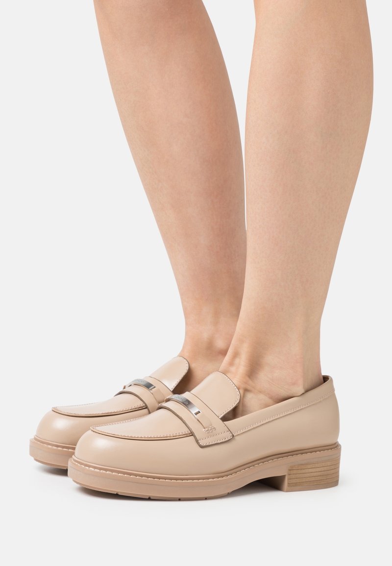 Mocassins en cuir beige avec bout rond, détails cousus et bande métallique accentuée. Talon épais et texturé pour une stabilité accrue.