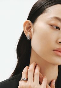 Pendientes de plata con una piedra ovalada central, rodeada por un diseño de estallido de sol texturizado; llevados con una chaqueta oscura y un anillo en la mano.