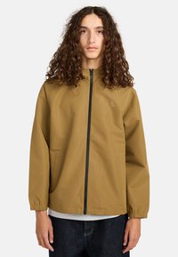 Junge Person mit langen lockigen Haaren, die eine beige Zip-Jacke, ein graues T-Shirt und dunkelblaue Jeans trägt und vor einem schlichten weißen Hintergrund steht.