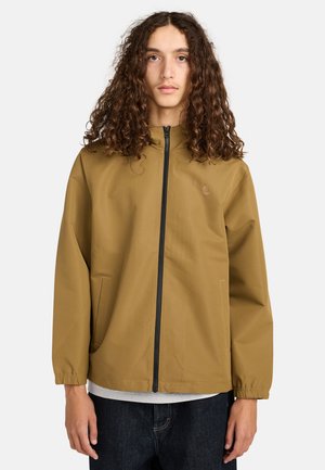 Jongpersoon met lang krullend haar, gekleed in een beige ritsjack, een grijze shirt en een donkere spijkerbroek, staand tegen een effen witte achtergrond.