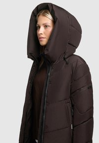 Brūns pūkains jopelis ar lielu kapuci. Tam ir rāvējslēdzēja priekšpuse, sānu kabatas un pārsteidzoša kviltota tekstūra. Gluda, matēta apdare.