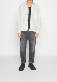 Giacca chiara, con bottoni, dotata di due tasche sul petto, indossata sopra una camicia nera, abbinata a jeans scuri, leggermente larghi, e scarpe nere.