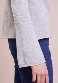 Seitliche Ansicht einer Person, die einen hellgrauen Pullover mit langen, weit geschnittenen Ärmeln und dunkelblauen Jeans trägt.