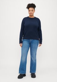 Merinovilla neulepusero, jossa on tekstuuri, pyöreä kaula-aukko ja punavalkoiset raidat hihoissa. Yhdistetty sinisiin leveälahkeisiin farkkuihin ja mustiin korkokenkiin.