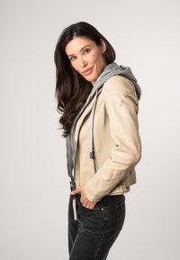 Beige leren jack met grijze capuchon, zilveren ritsaccenten en zijzakken, gedragen over een grijs shirt en zwarte jeans. Glad textuur.