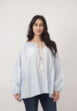 Kobieta z długimi ciemnymi włosami, ubrana w luźną jasnoniebieską bluzkę z frędzlami i ciemne dżinsy, stojąca na tle jednolitego jasnego tła.