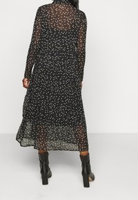 L�ång, svart, genomskinlig klänning med lagerdesign, framhävd av ett spritt mönster av vita och guldiga löv. Parat med svarta klackstövlar.