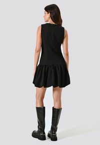 Vestito nero senza maniche con un busto aderente, orlo a campana con volant e zip sul retro. Un paio di stivali in pelle nera al ginocchio completa il look.