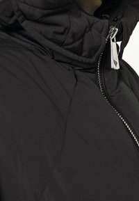 Černá prošívaná péřovka s vysokým límcem, která má stříbrný zip a hladký povrch. Žádné další vzory ani hardware nejsou viditelné.