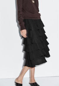 Black midi skirt with layered ruffles. Made of lightweight fabric, it features a textured finish and a flowing design. Paired with a brown sweater.