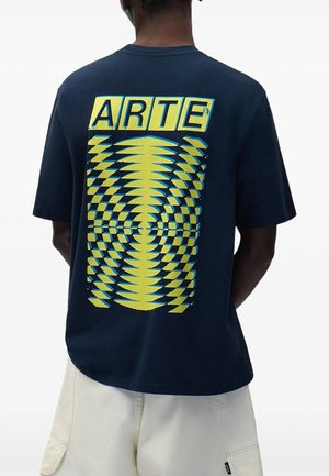 Persona che indossa una maglietta blu navy con motivo a scacchi giallo distorto e la parola "ARTE" sulla schiena, abbinata a pantaloncini di colore chiaro.